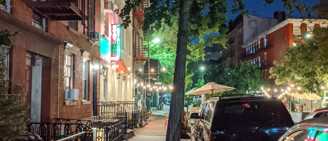 Neon-lit Manhattan street scene at night with illuminated diner signage, yellow cabs, and night owls walking along Greenwich Village